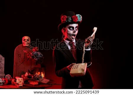 Goddess of death woman using landline phone in studio, celebrating la cavalera catrina or santa muerte on traditional mexican holiday. Holding cord telephone dressed for day of the dead.
