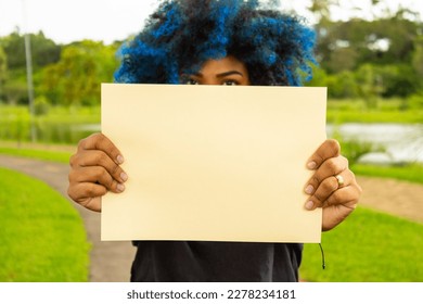 GOIANIA GOIAS BRAZIL - MARCH 20 2023: A young woman, with hair dyed blue, her face hidden behind a blank poster, with a landscape in the background.