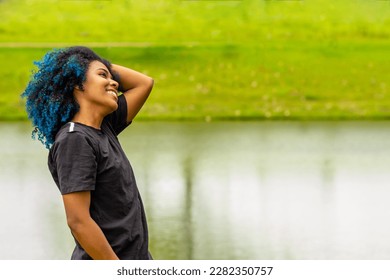 GOIANIA GOIAS BRAZIL - MARCH 20 2023: A young black woman, happy, in a very green natural park, straightening her dyed blue hair.