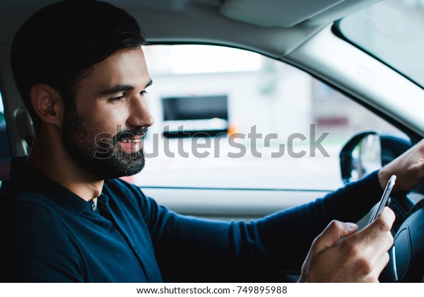 Going for a date. Side\
view of handsome young man using his smart phone with smile\
while\
sitting in car\
