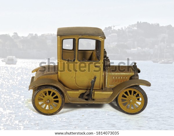 Gold antique model car in\
overlay with blurry lake and mountain village background​\
reflecting the sun
