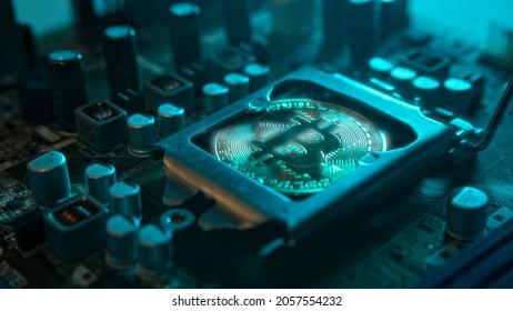 A gold Bitcoin coin with glare and reflection installed instead of a central processor in the motherboard with electronic components with blue light and smoke in the darkness. Bitcoin Mining - Shutterstock ID 2057554232