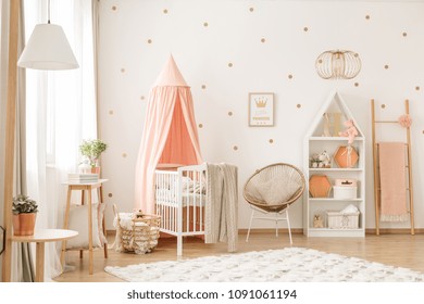 Gold chair with pillow standing next to a white wooden crib with pastel pink canopy in bright baby room interior with dots on the wall