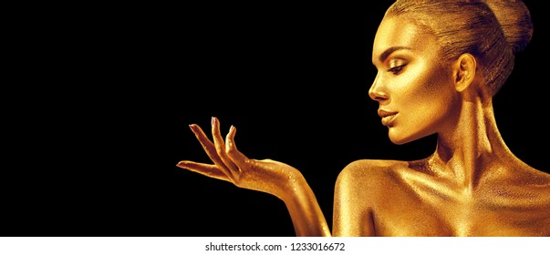 Gold Christmas Woman. Beauty fashion model girl with Golden make up, hair and jewellery, pointing hand on black background. Gold glowing skin. Metallic, glance Fashion art portrait, Hairstyle, make up