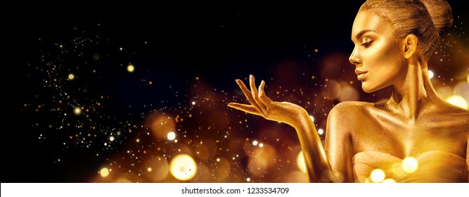 Gold Christmas Woman. Beauty fashion model girl with Golden make up, hair and jewellery, pointing hand on black background. Gold glowing skin. Metallic, glance Fashion art portrait, Hairstyle, make up