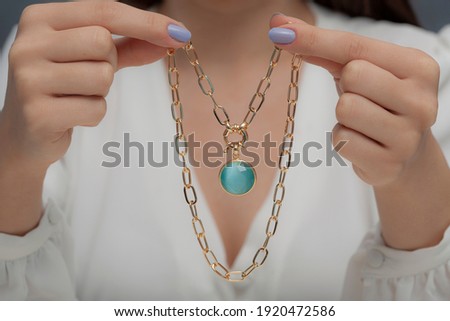 Gold necklace with blue stone on hand of lady with blue nail polish in white clothes.