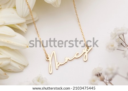 Gold necklace with name inscription displayed on a floral background. Jewelry photo for e-commerce, online sale, social media.