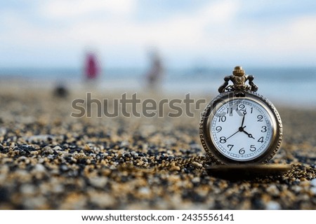 Gold pocket watch lies on a sandy beach with a person walking on the beach in the background. With copy space for text