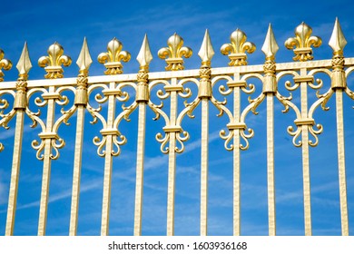 Gold railings with intricate fleur di lis designs shining in the sunlight against a blue sky outside the gates of Versailles, France