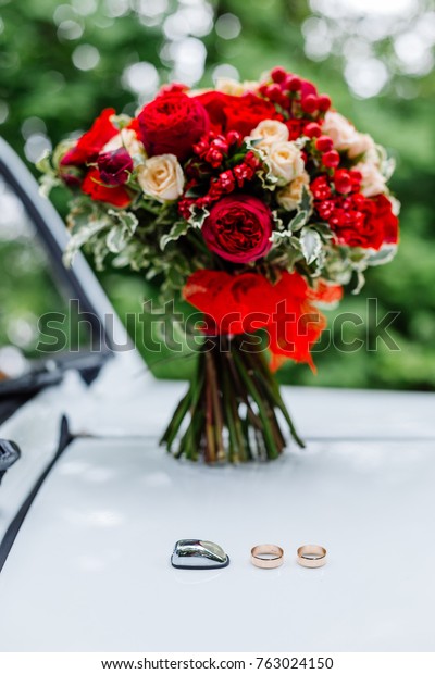 Gold rings lying on white car in\
background of passion wedding bouquet with dark red and marsala\
roses, greenery. Bridal flowers, decorated with\
ribbon