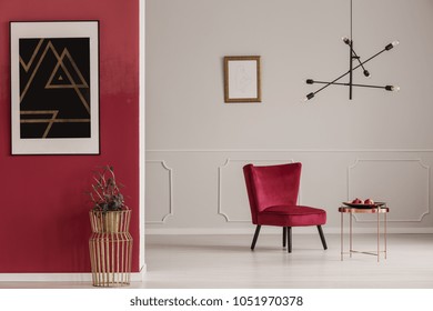 Gold Table Against Red Wall With Black Poster In Apartment Interior With Armchair Against White Wall