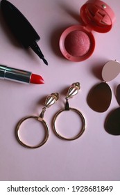 Gold vintage earrings, red lipstick, eyeliner and blush on pink background. Top view.