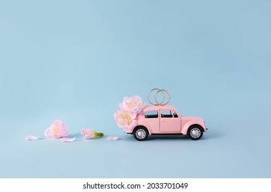 Gold wedding rings on miniature toy retro car decorated with pink flowers on blue background. The concept of marriage. Copy space.