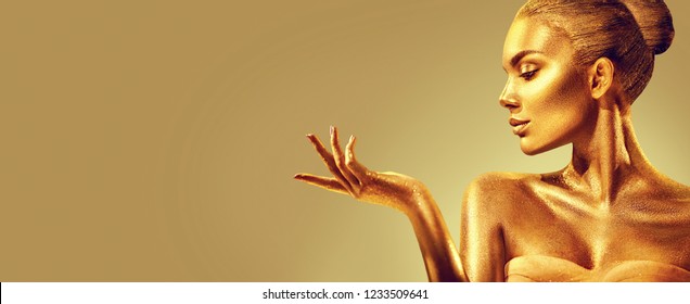 Gold Woman Skin. Christmas Beauty Fashion Model Girl With Golden Make-up, Hair And Jewellery On Gold Background. Pointing Hand, Proposing Products, Sales. Metallic Glance Fashion Art Portrait, Glamour