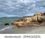 Golden big boulders sunbathed by the late afternoon sun. Brown and orange stones, green turquoise sea, sky full of gray and white clouds, mountains in the horizon. Jurere Internacional beach.