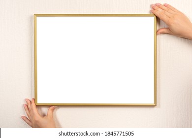 Golden Blank Picture Frame Hanging On A Beige Wall. Hands Hanging Photo Frame Mockup On Wall. Young Woman Holding Empty Picture Frame With Copyspace