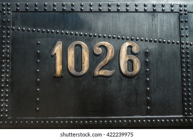 Golden Colored Copper Number 1026 On Black Riveted Metal Surface Of Aged Vintage Steam Locomotive 