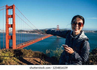 Golden Gate Bridge Cheerful Asian Girl Tourist Sightseeing In San Francisco USA. Young Woman Tour Enjoying The View At The Famous Travel Landmark In California USA Pointing Finger And Using Cellphone