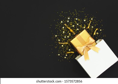 Golden Gift With Ribbon From White Paper Bag, Confetti Stars On Black Background. Flat Lay, Top View, Copy Space. Golden Decorations, Party, Birthday, Holiday Concept. New Year Christmas Composition