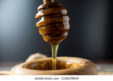 Golden honey flows from the stick into a wooden jar. Aromatic nectar dripping from honey-dipper. Honey in a bowl. Dripping honey from wooden dipper on wooden background. Selective focus.