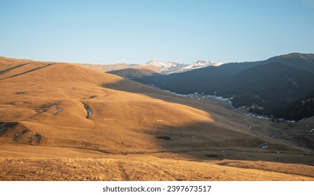 Golden hour light bathes rolling hills and a sparse mountain valley, leading to snow-dusted peaks and dark coniferous forests under a clear evening sky. - Powered by Shutterstock