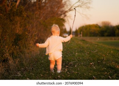 Golden Hour Photo Of Young Toddler Dressed In White Happily Playing With A Stick Outside. Carefree, Happy, One Year Old Boy.