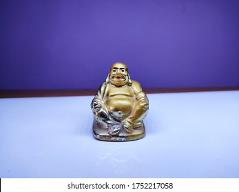 A golden Laughing Buddha figurine sits on a white surface with a purple backdrop, showcasing its joyful pose, smiling face, and plump figure, symbolizing happiness, luck, and prosperity. - Powered by Shutterstock