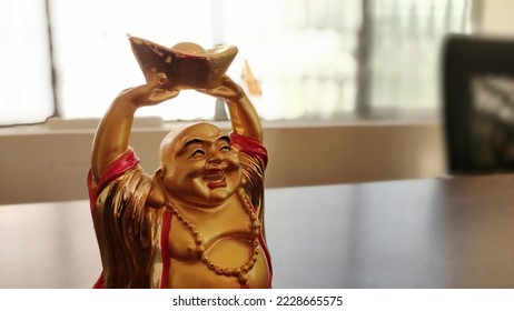 A golden Laughing Buddha statue holding a gold ingot with his hands upright. The statue is smiling broadly and wearing a red robe with a beaded necklace. - Powered by Shutterstock