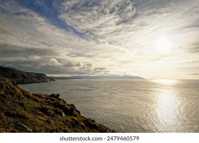 The golden light of sunset illuminates the ocean and coastal cliffs under a cloud-covered sky - Powered by Shutterstock