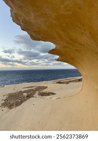 Golden limestone cave frames dramatic Mediterranean seascape at sunset in Gozo, Malta. Curved rock formation creates natural window, revealing deep blue waters, sandy beach, and cloudy horizon beneath