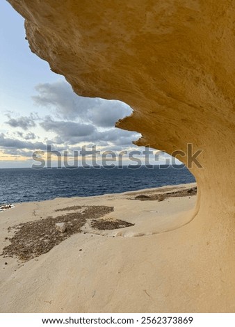 Golden limestone cave frames dramatic Mediterranean seascape at sunset in Gozo, Malta. Curved rock formation creates natural window, revealing deep blue waters, sandy beach, and cloudy horizon beneath