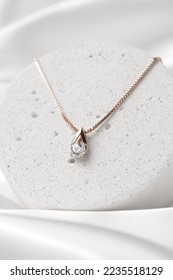 Golden necklace with crystals on concrete podium on white silk background, rose gold, single diamond pendant. Beautiful accessories for women. Elegant jewelery gift or present for wedding or saint