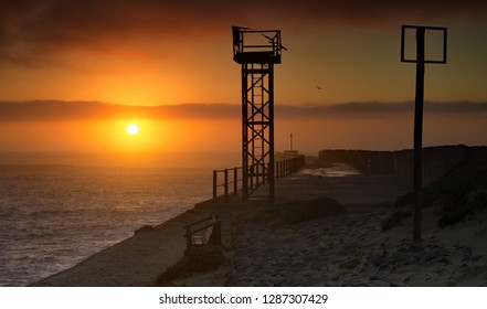 The Golden Orb Of The Sun Breaks The Horizon At Dawn, Between The Indian Ocean And A Band Of Dark Cloud. In The Foreground, A Rusting Tower Is Silhouetted On A Breakwater At The Mouth Of A River.