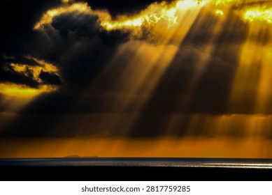 Golden rays of light break through stormy clouds, illuminating the sea and a distant island under a vibrant sunset sky
