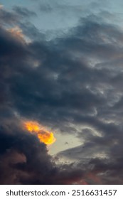 Golden rays of setting sun light give the impression of a fire breathing dragon peering out from amongst storm clouds gathering in front of a pale blue summer evening sky