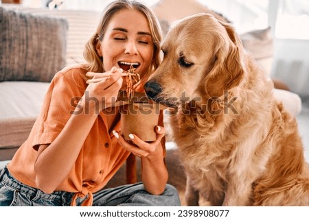 Golden retriever eagerly licking paper box with chinese noodles being eating by young woman. Female pet owner sharing food with lovely furry friend at cozy apartment.                               