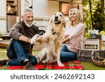 Golden retriever shaking his paw with aged handsome smiling man while his wife watching them on blanket near travel car. Mature travelers caravanning with pet dog together