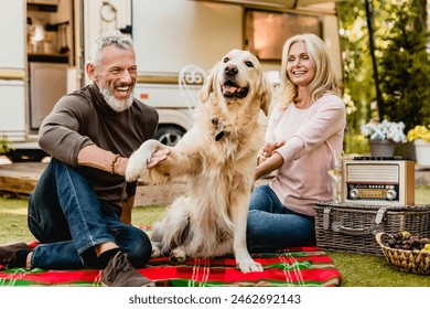 Golden retriever shaking his paw with aged handsome smiling man while his wife watching them on blanket near travel car. Mature travelers caravanning with pet dog together - Powered by Shutterstock