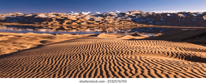 Golden ripples of sand stretch across a sunlit desert. Snow-capped peaks in the distance reflect in a shimmering blue lake beneath a clear sky. - Powered by Shutterstock