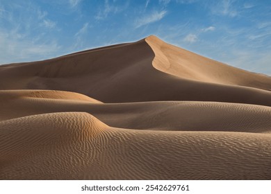 Golden sand dune in Le Khwair, Oman, surrounded by smooth, rippling sand patterns, under a bright blue sky with scattered clouds. - Powered by Shutterstock