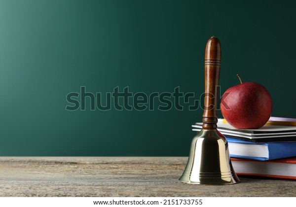 Golden school bell, apple and books on\
wooden table near green chalkboard. Space for\
text