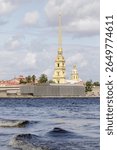Golden spire of Peter and Paul Cathedral rises above fortress walls, viewed from across the Neva River under cloudy summer skies.