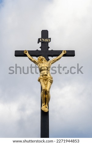 Golden statue of crucified Jesus with a Latin inscription INRI, standing on an iron cross, against a cloudy background.