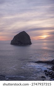 Golden sun dips behind Haystack Rock at Cape Kiwanda on the Pacific coast of Oregon, USA. Lonely sea stack stands bold against colorful sky. Waves gently break on the rocky shore. Sky paints warm hues of orange, purple, and lavender