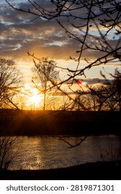 Golden sun rays piercing through silhouetted tree branches over a tranquil river at sunset, creating a serene atmosphere with dramatic clouds in the warm evening sky