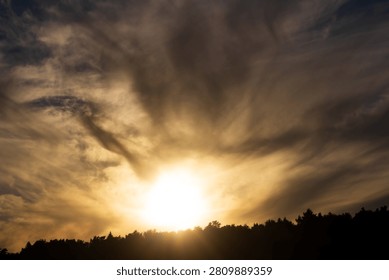 Golden sunbeams breaking through dramatic, swirling storm clouds above a dark forest silhouette, creating a divine and moody celestial display of natural beauty during late afternoon golden hour