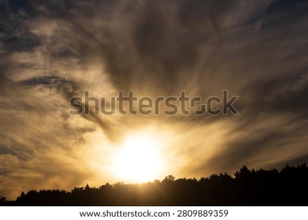 Golden sunbeams breaking through dramatic, swirling storm clouds above a dark forest silhouette, creating a divine and moody celestial display of natural beauty during late afternoon golden hour