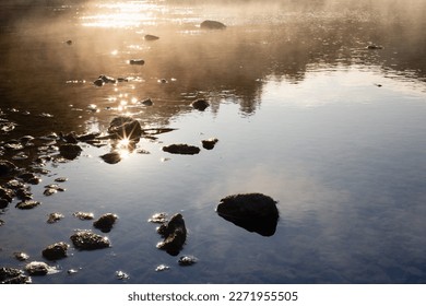 Golden sunbeams and ripple glare on calm river water surface with fluffy yellow fog, blue reflection of sky, stones on shore in early morning. Bright, warm, quiet sunrise background, detail, texture. - Powered by Shutterstock