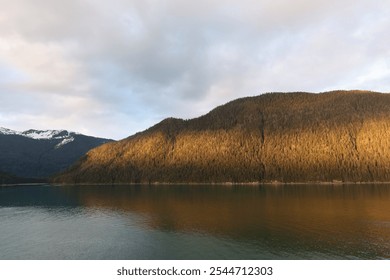 Golden sunlight illuminates forested mountain slopes beside calm waters near Juneau, Alaska. Snow-capped peaks reflect in turquoise glacial waters under moody clouds. - Powered by Shutterstock