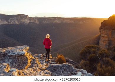 Golden sunlight streaming into valley with rising sandstone escarpment mountain and onto a cliff ledge where a female hiker standing taking in the magnificent views - Powered by Shutterstock